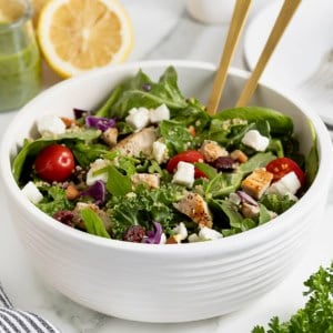 A green salad with tomato, feta, and quinoa in a large white bowl with two gold tone spoon handles sticking out.