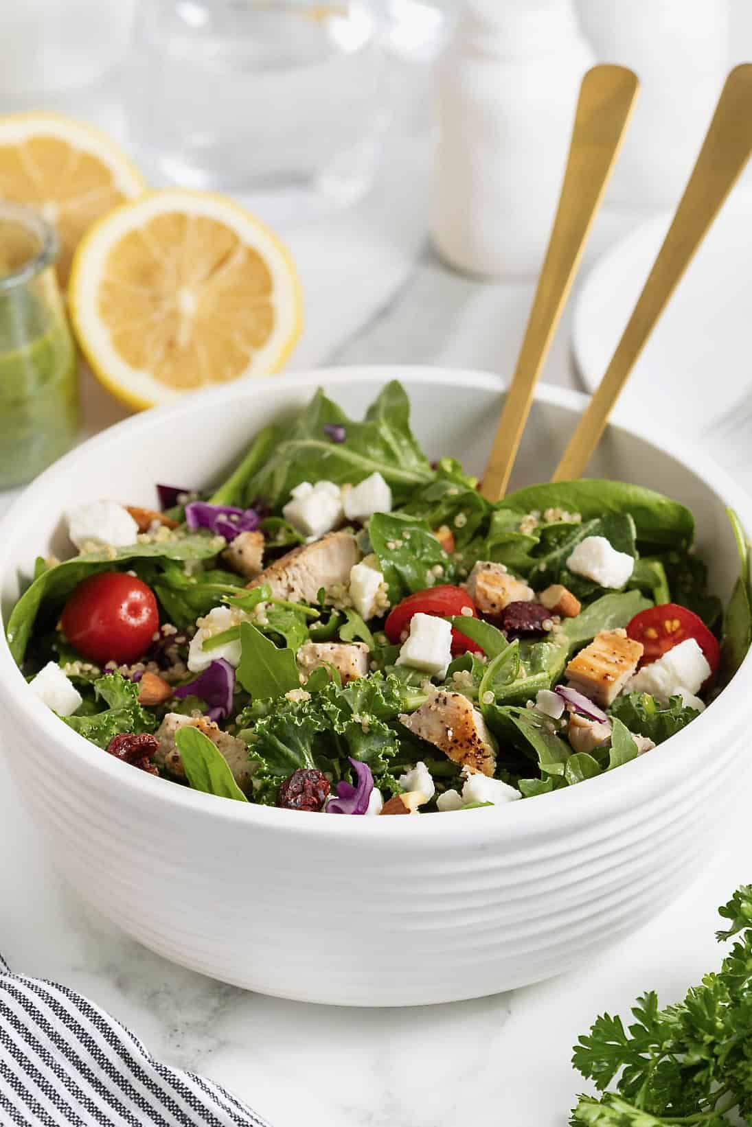 A green salad with tomato, feta, and quinoa in a large white bowl with two wooden spoon handles sticking out.