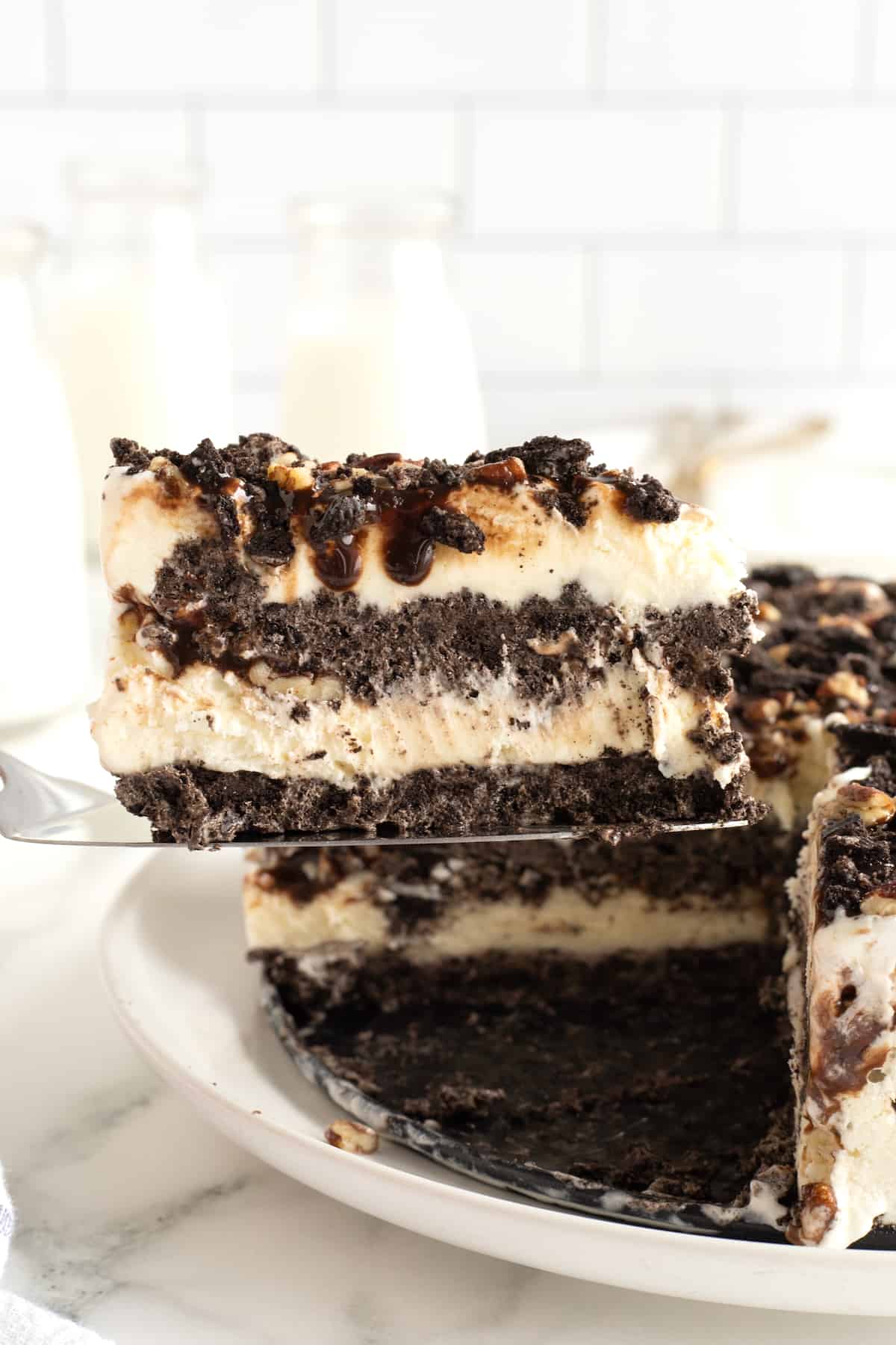 A slice of ice cream pie with an Oreo crust, ice ream filling, and crushed Oreos, chopped pecans and chocolate syrup on top.