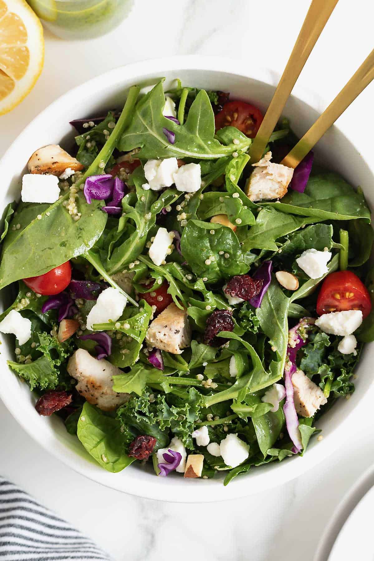 A green salad with tomato, feta, and quinoa in a large white bowl with two golden spoon handles sticking out.