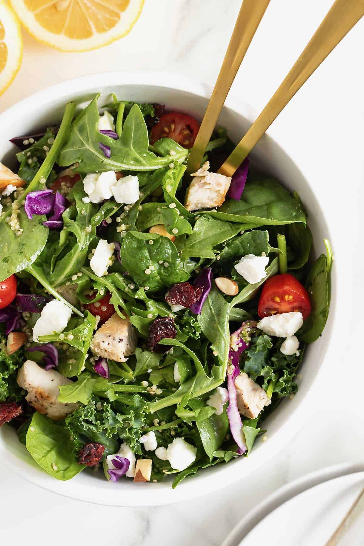 A green salad with tomato, feta, and quinoa in a large white bowl with two golden spoon handles sticking out.