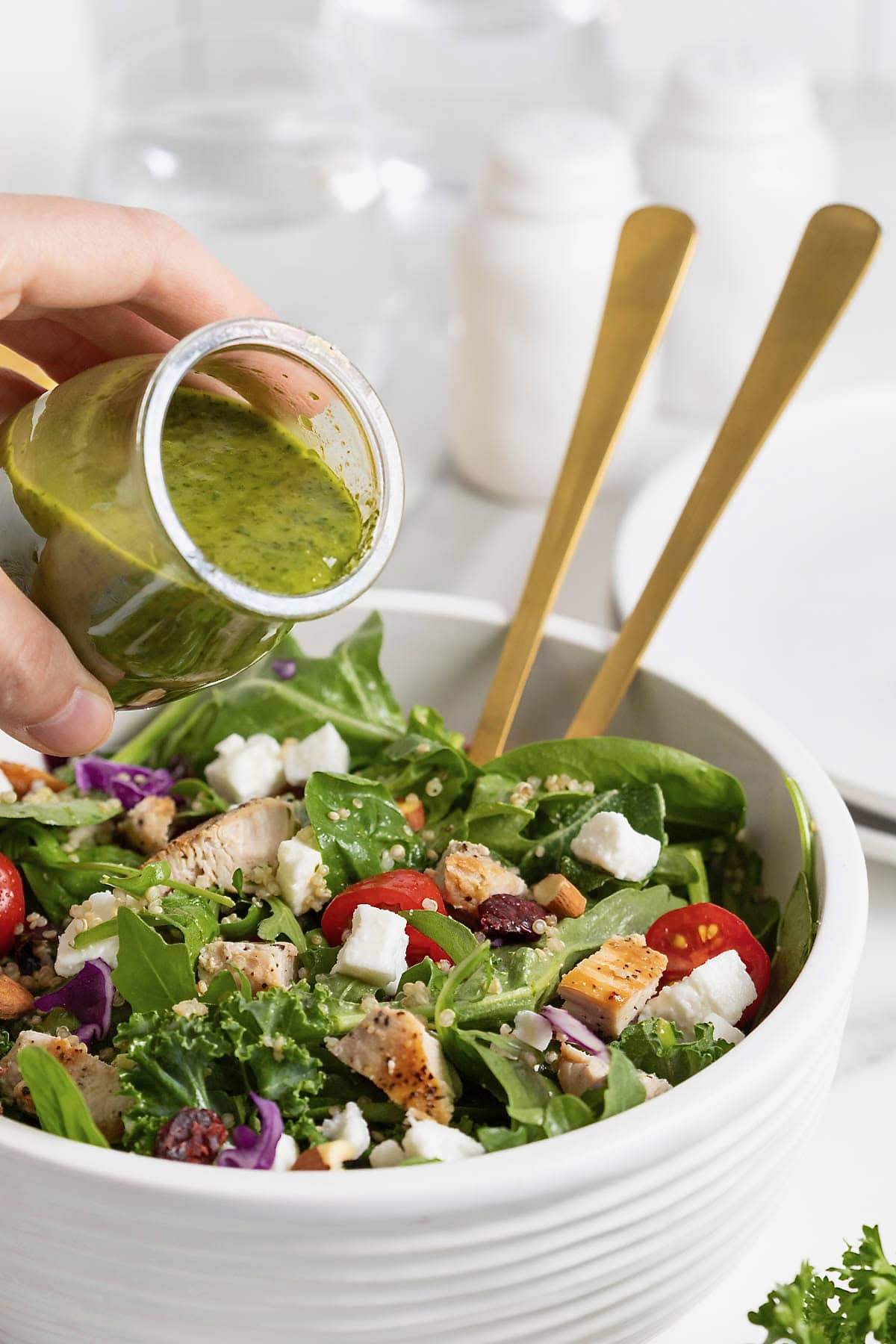 Green salad dressing being poured from a small glass carafe onto a green salad in a large white bowl.