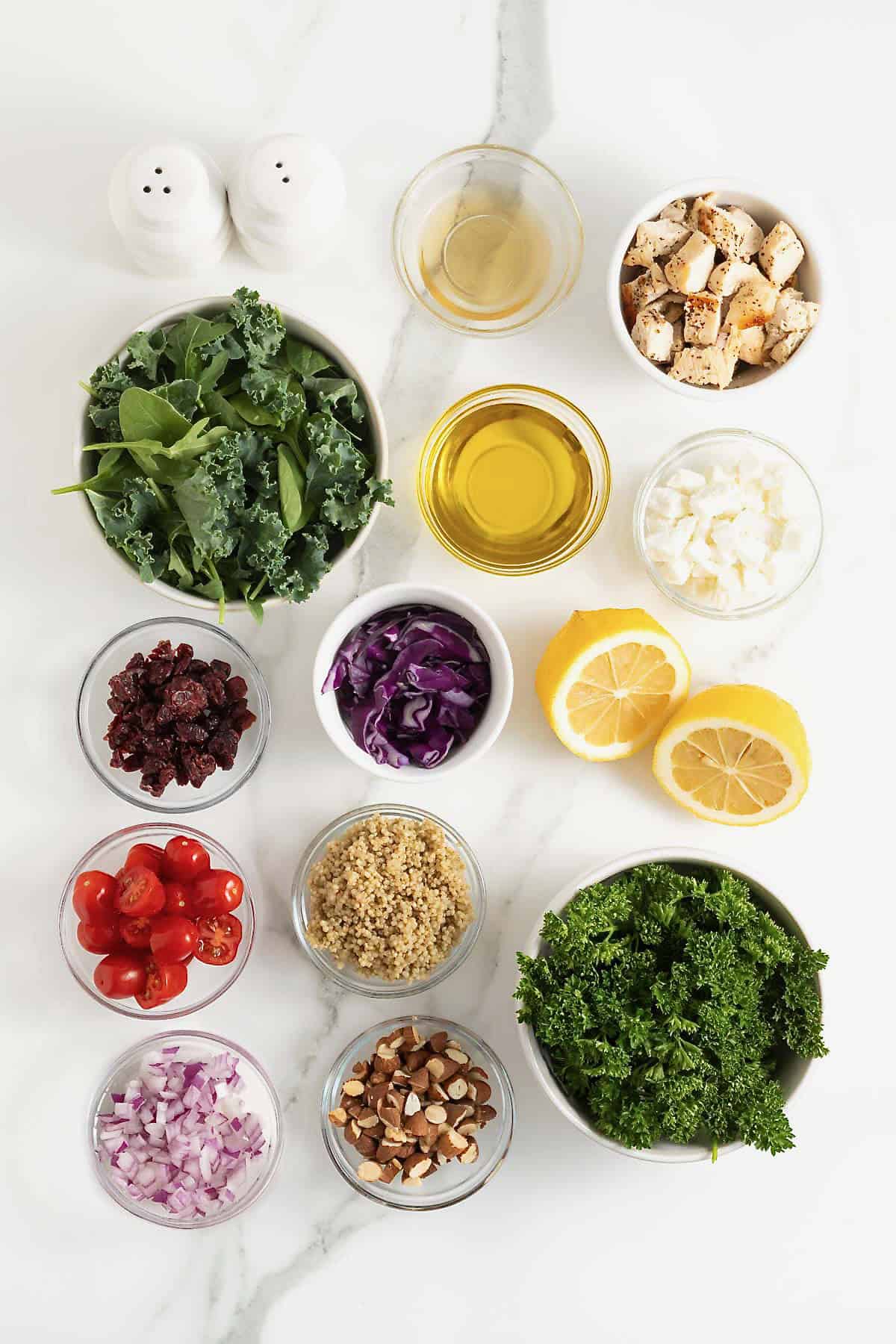 Ingredients to make a chicken quinoa salad in small glass dishes on a white marble counter.