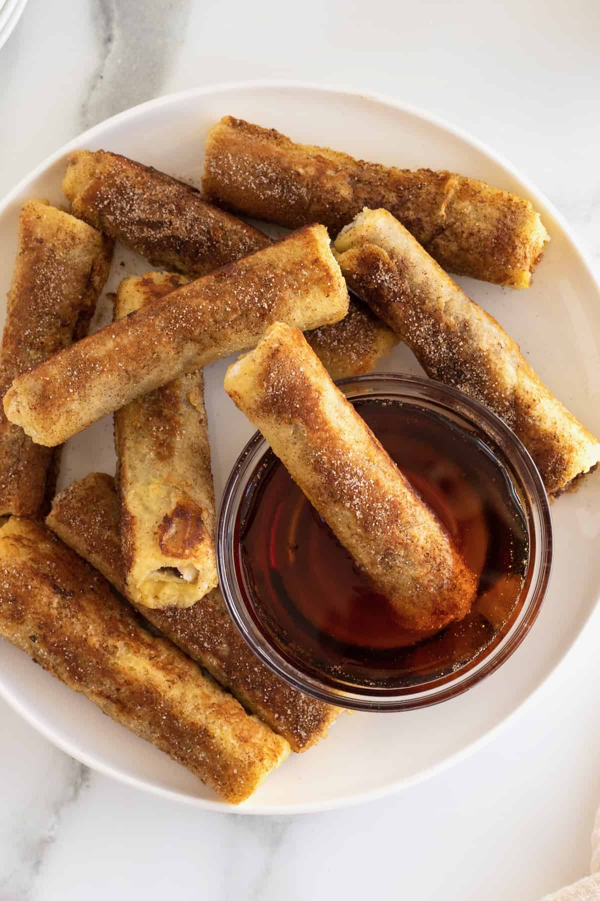 Cinnamon sugar covered French toast roll ups surrounding a glass bowl of warmed maple syrup.