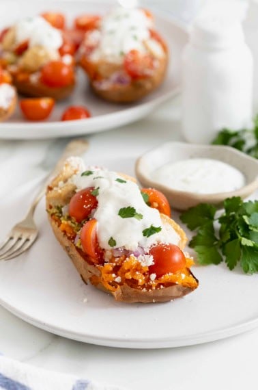 A halved sweet potato topped with hummus, tabouleh, chicken, tomatoes, red onion, feta, and tzatziki sauce on a white plate.
