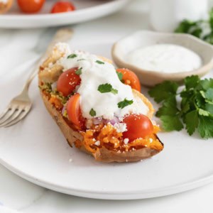 A halved sweet potato topped with hummus, tabouleh, chicken, tomatoes, red onion, feta, and tzatziki sauce on a white plate.