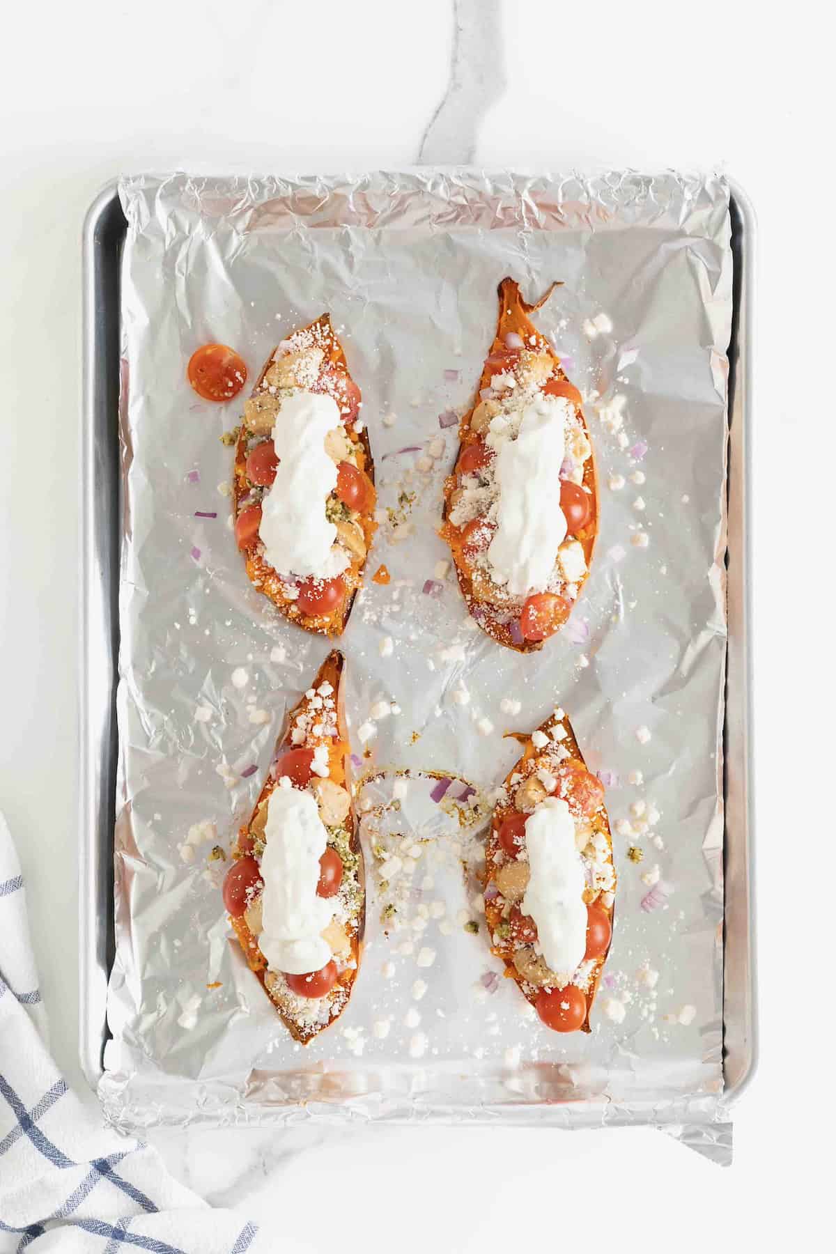 Four sweet potato halves topped with hummus, tabouleh, tomatoes, red onion, feta, and greek yogurt on a foil lined baking sheet.