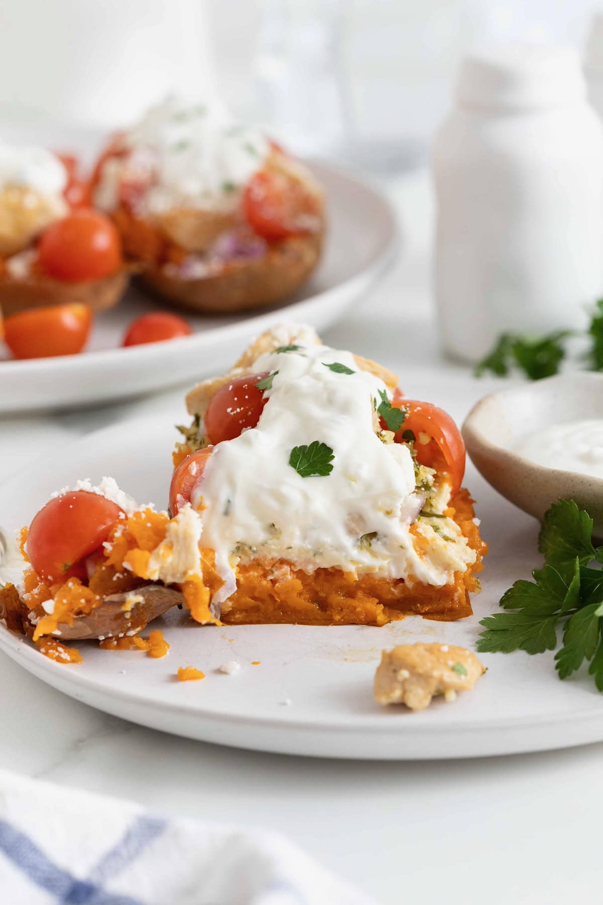 A bite taken out of a halved sweet potato topped with hummus, tabouleh, chicken, tomatoes, red onion, feta, and tzatziki sauce on a white plate.