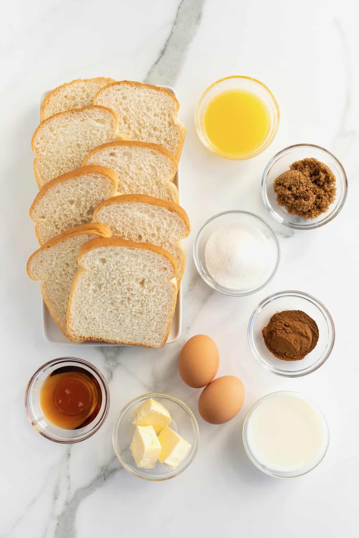 Sliced white bread on a counter next to two brown eggs and glass dishes filled with maple syrup, cinnamon, sugar, and melted butter.