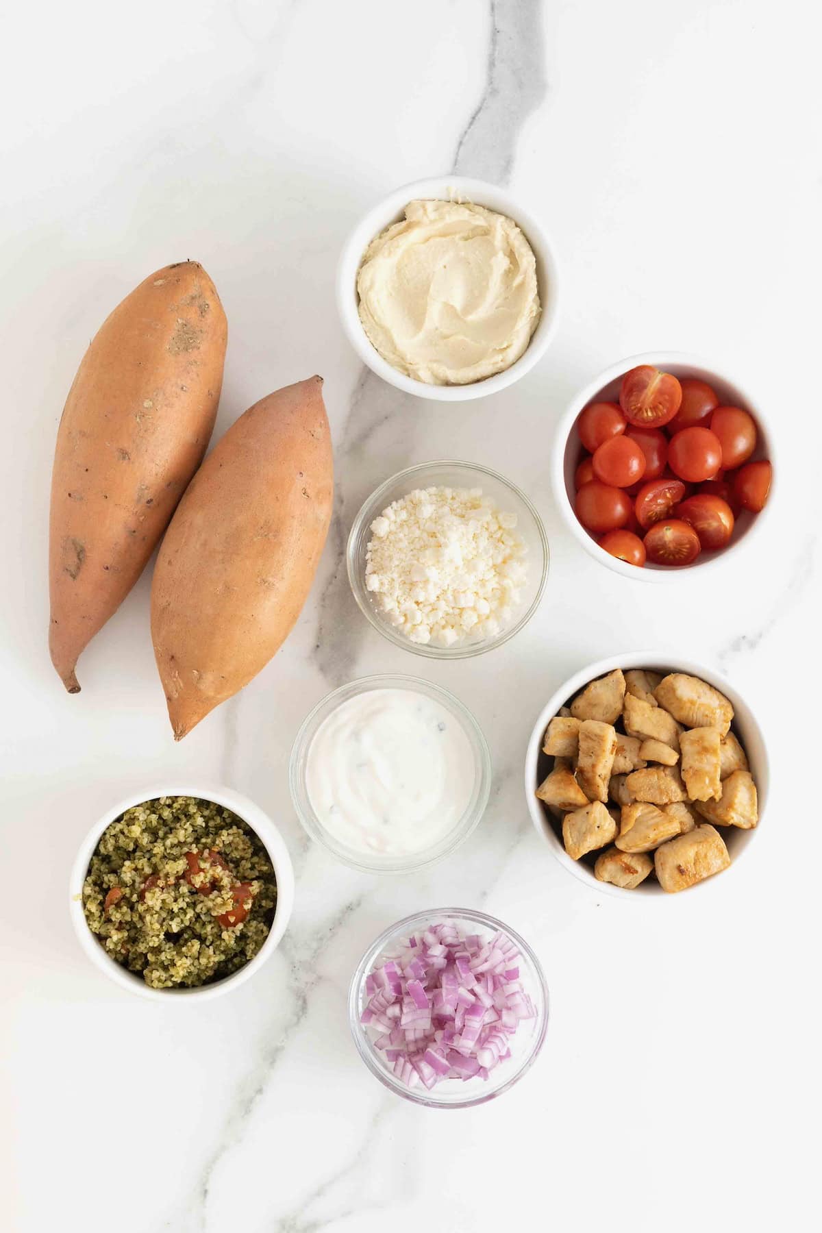 Ingredients to make Greek Loaded Sweet Potatoes in small white dishes on a white marble counter.