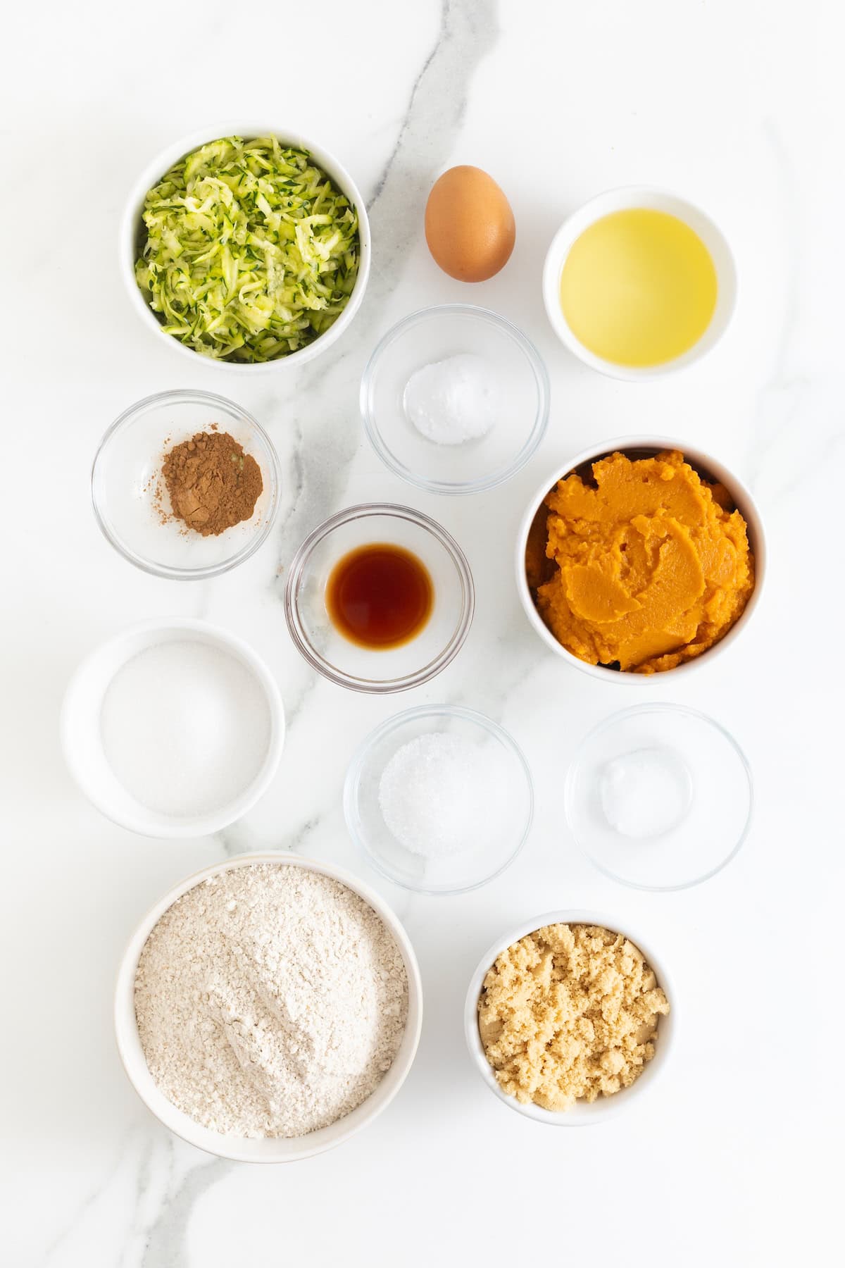 Ingredients to make whole wheat pumpkin zucchini bread in small glass dishes on a white marble counter.