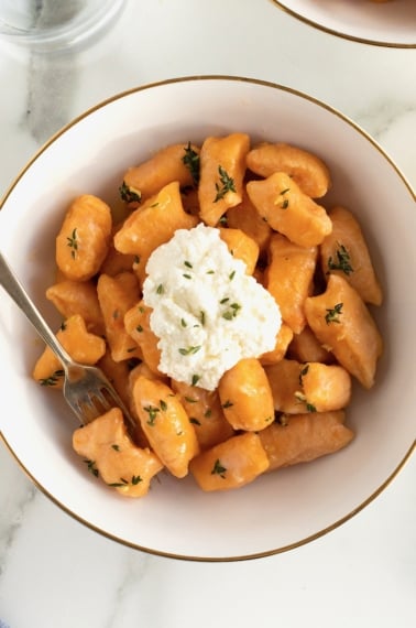 A gold-rimmed white bowl of sweet potato gnocchi with a dollop of fresh ricotta on top.