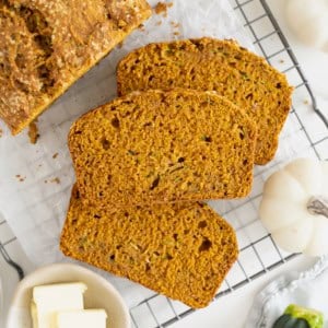 Three slices of whole wheat pumpkin zucchini bread next to the loaf on a parchment lined cooling rack.