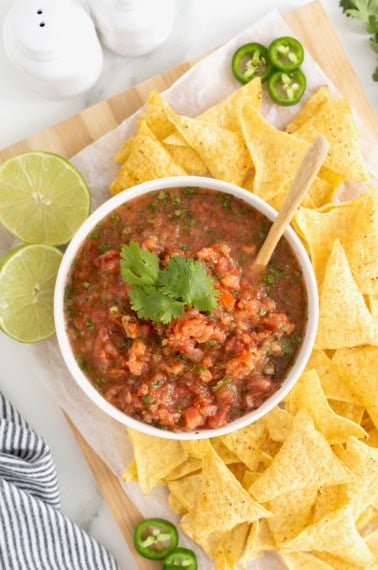 A white bowl of salsa on a wooden cutting board surrounded by corn chips.