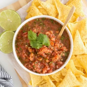 A white bowl of salsa on a wooden cutting board surrounded by corn chips.