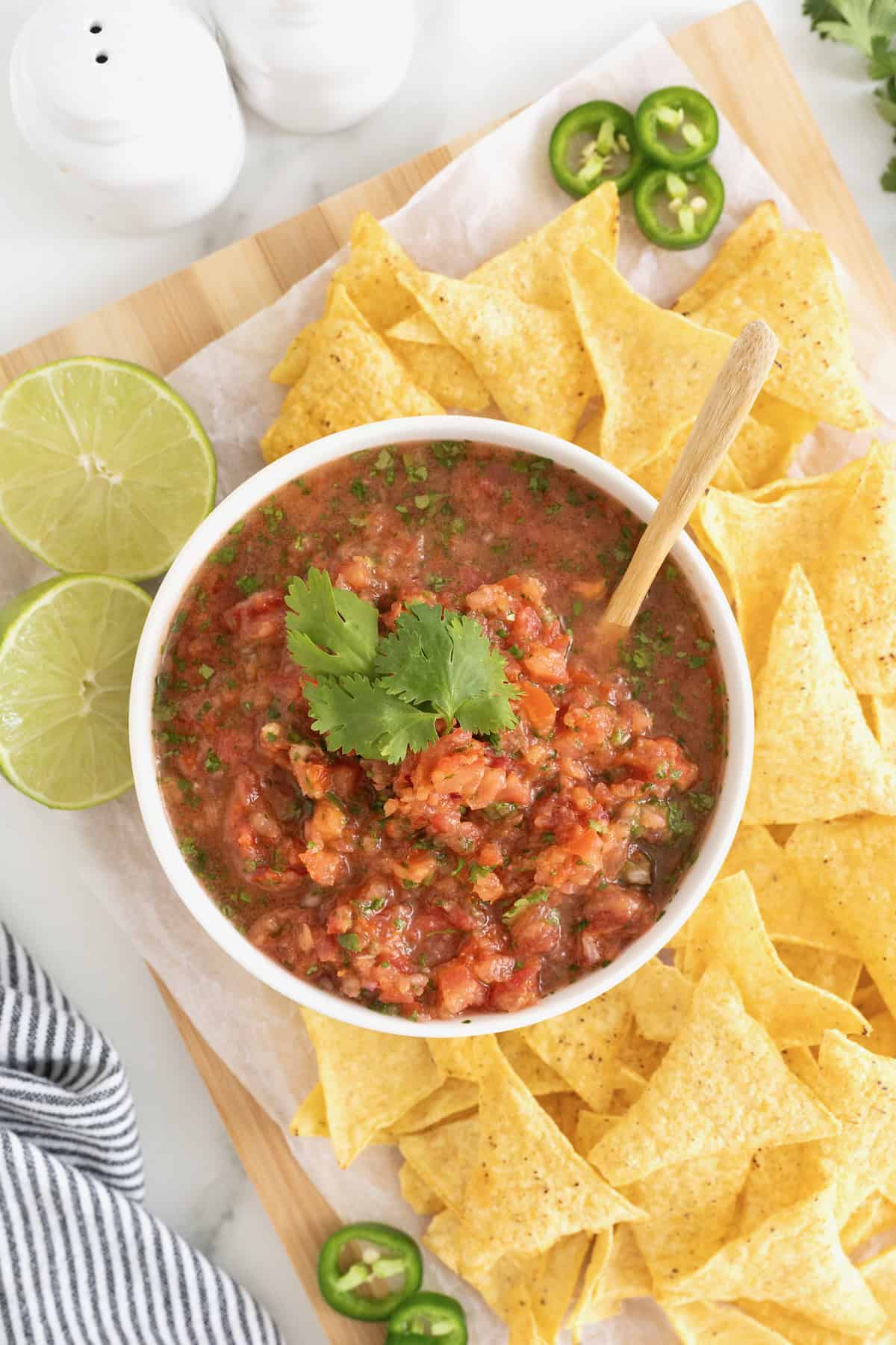A white bowl of salsa on a wooden cutting board surrounded by corn chips.