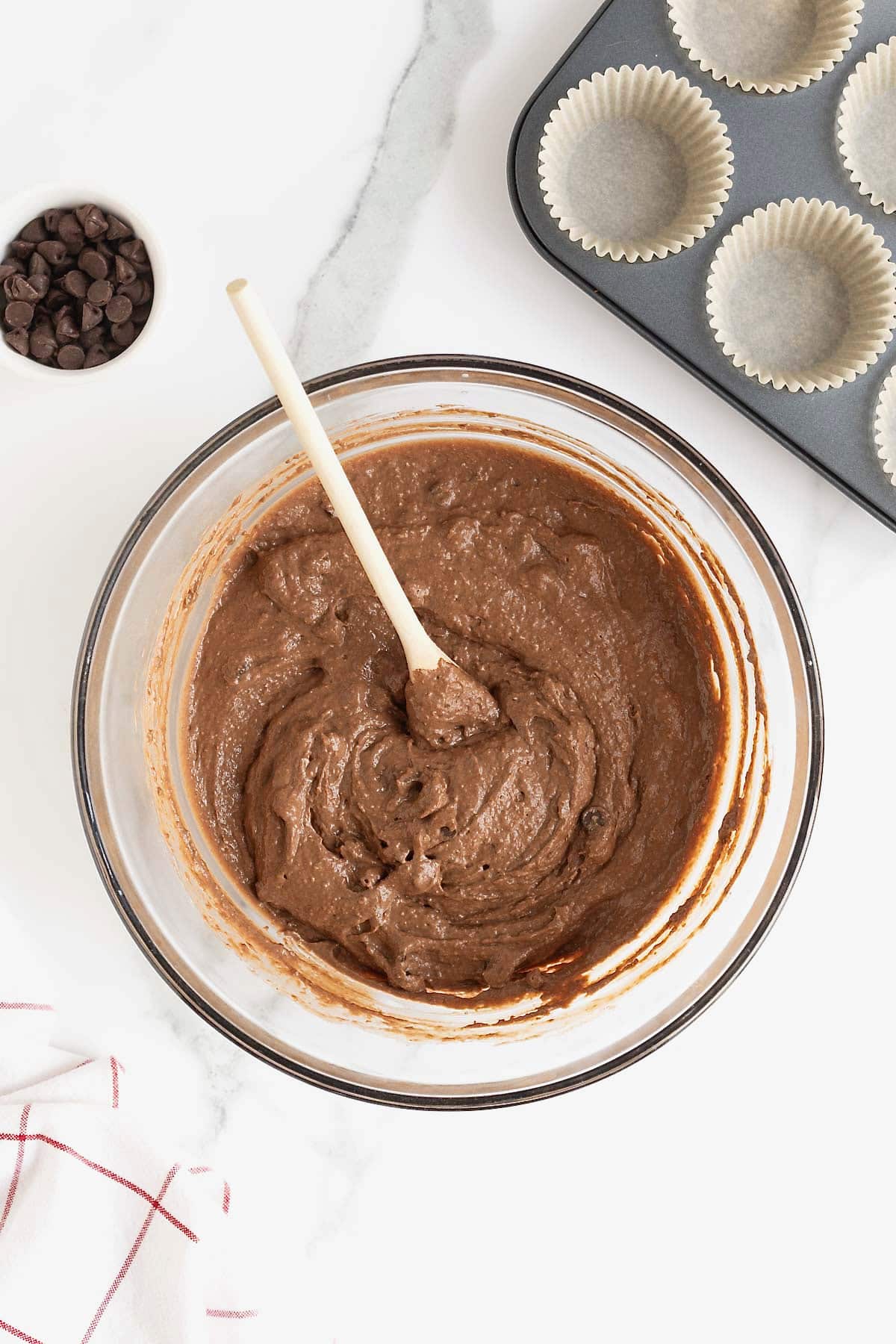 Chocolate muffin batter in a glass mixing bowl with a wooden spoon sticking out.
