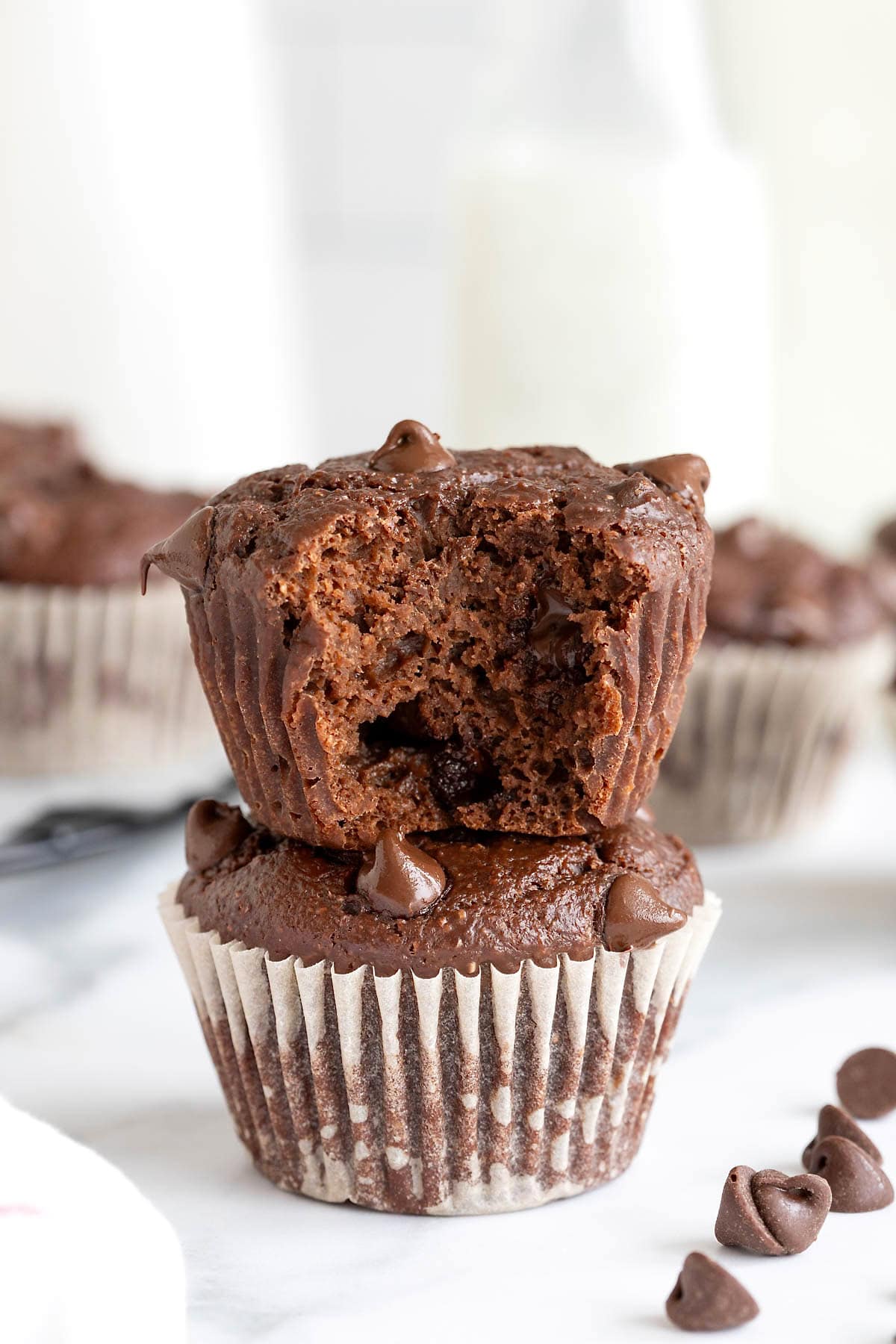 Two chocolate muffins with white paper wrappers stacked on a white marble counter. The top muffin has a bite out of it.