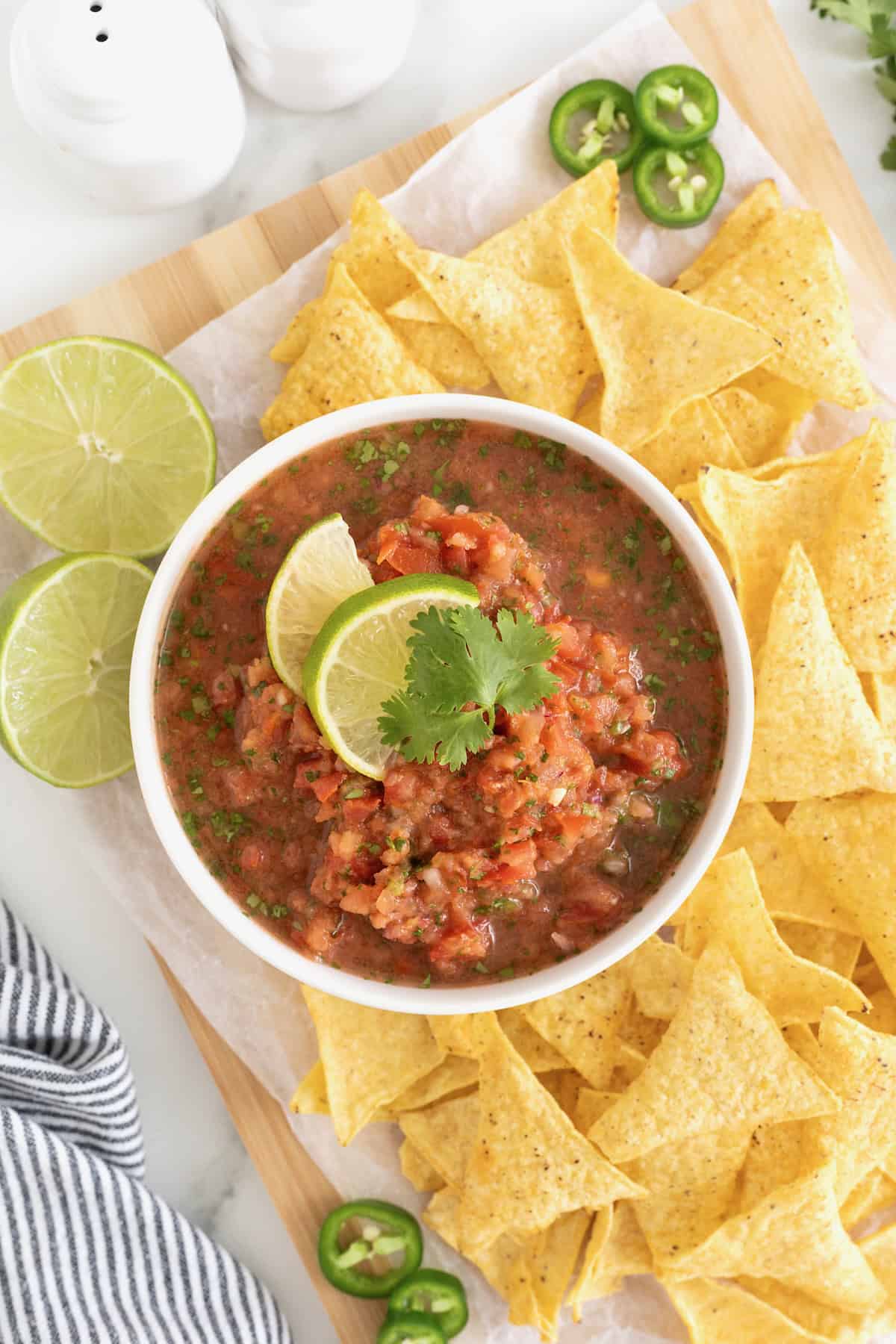 A white bowl of salsa on a wooden cutting board surrounded by corn chips.