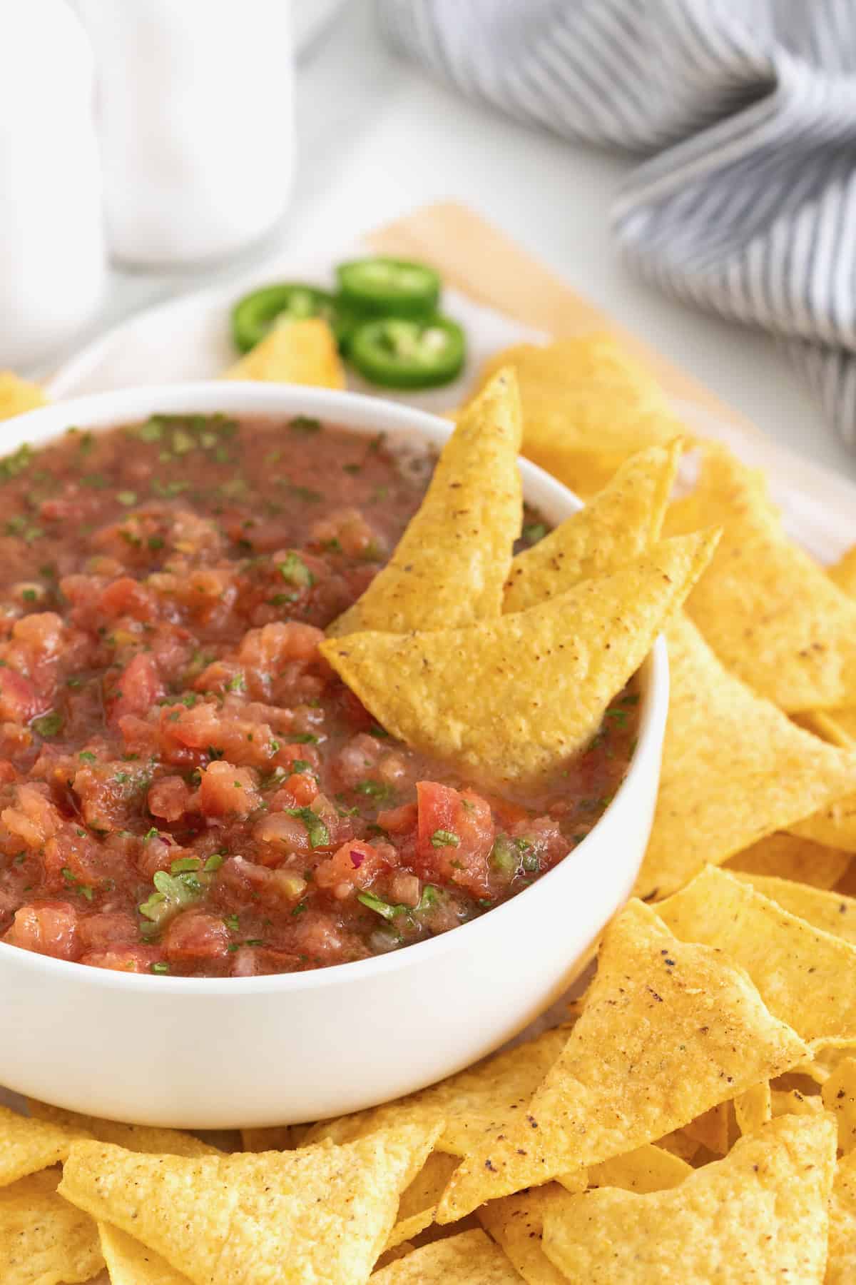 A white serving bowl of salsa with three tortilla chips sticking out.