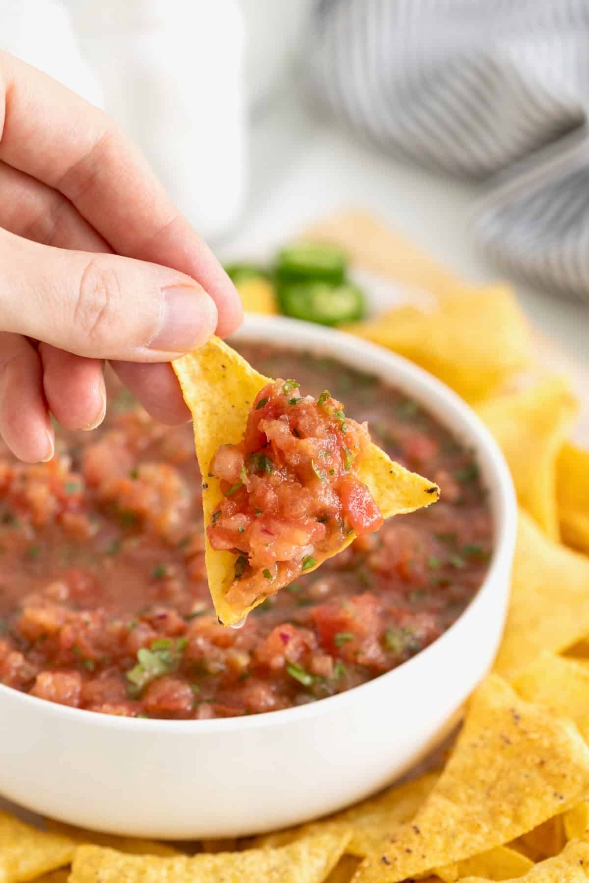 A hand holding a corn chip with salsa on it. In the background is a white serving bowl of homemade salsa surrounded by corn chips.
