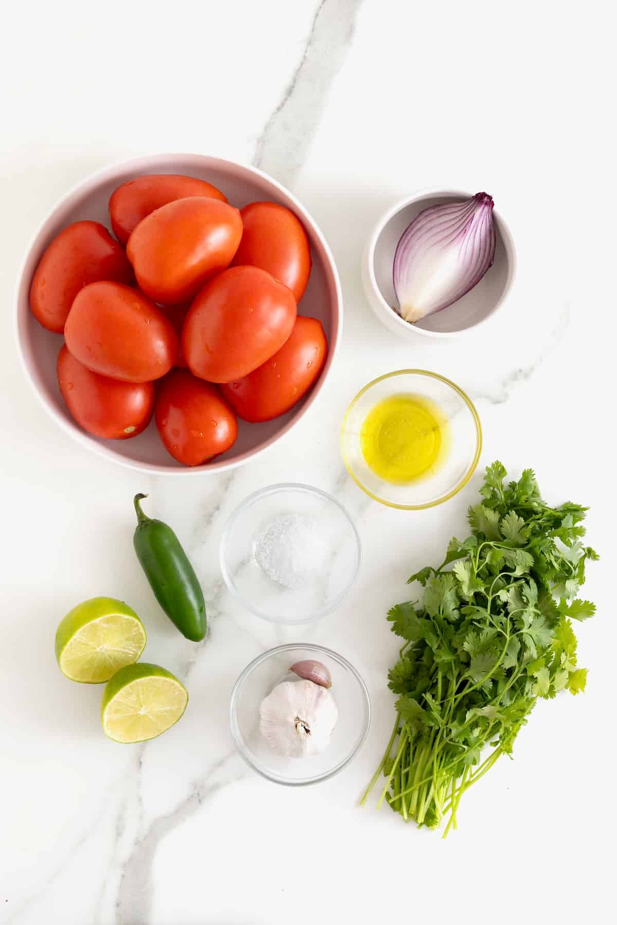 Ingredients to make homemade salsa in glass dishes on a white marble counter.
