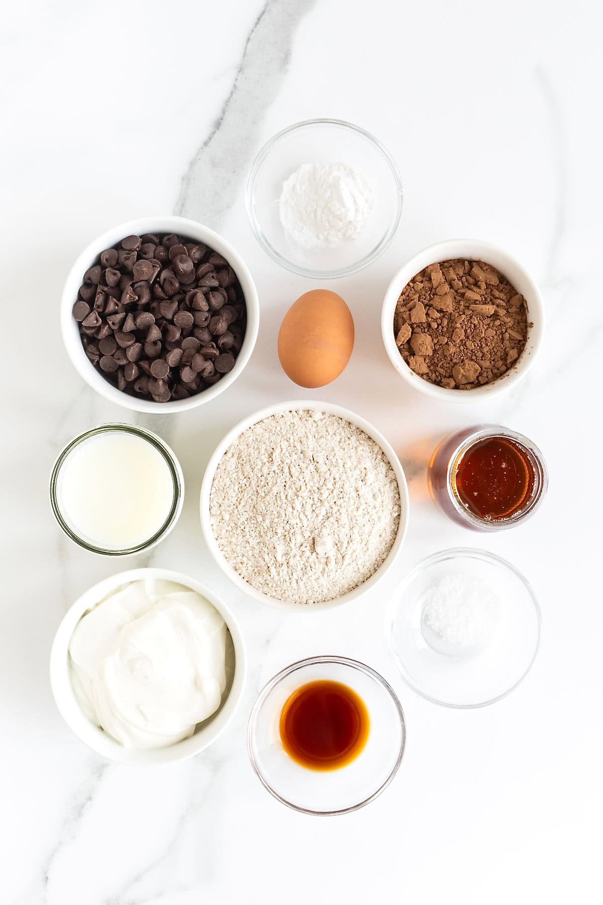 Ingredients to make healthier double chocolate muffins in small white dishes on a white marble counter.