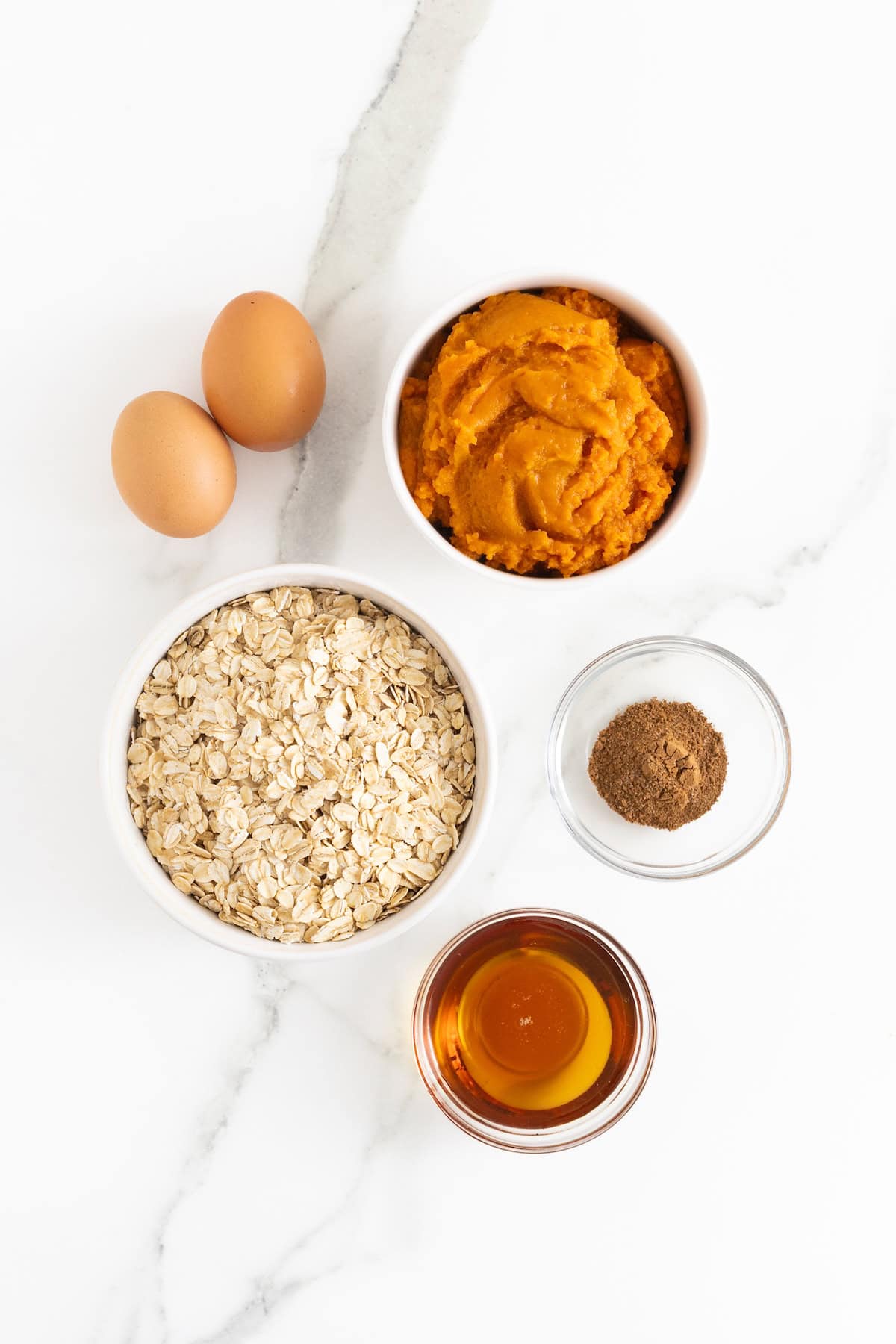 Ingredients to make pumpkin oatmeal pancakes in small glass dishes on a white marble counter.