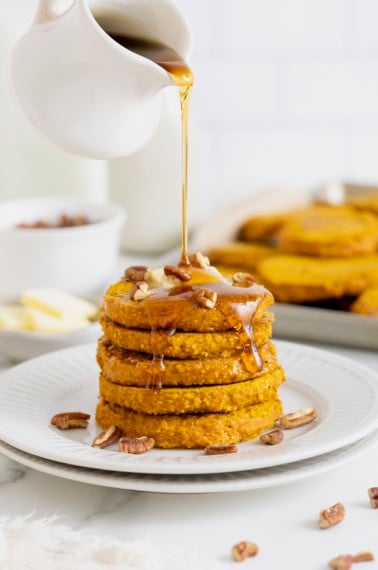 A stack of five pumpkin oatmeal pancakes on a white plate. Syrup is being drizzled onto the pancakes from a white pitcher.