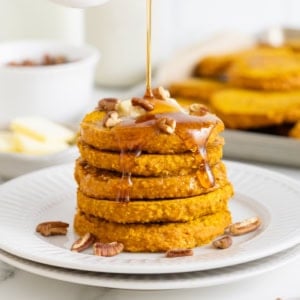 A stack of five pumpkin oatmeal pancakes on a white plate. Syrup is being drizzled onto the pancakes from a white pitcher.