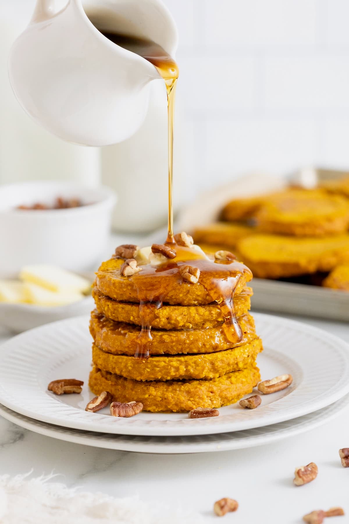 A stack of five pumpkin oatmeal pancakes on a white plate. Syrup is being drizzled onto the pancakes from a white pitcher.