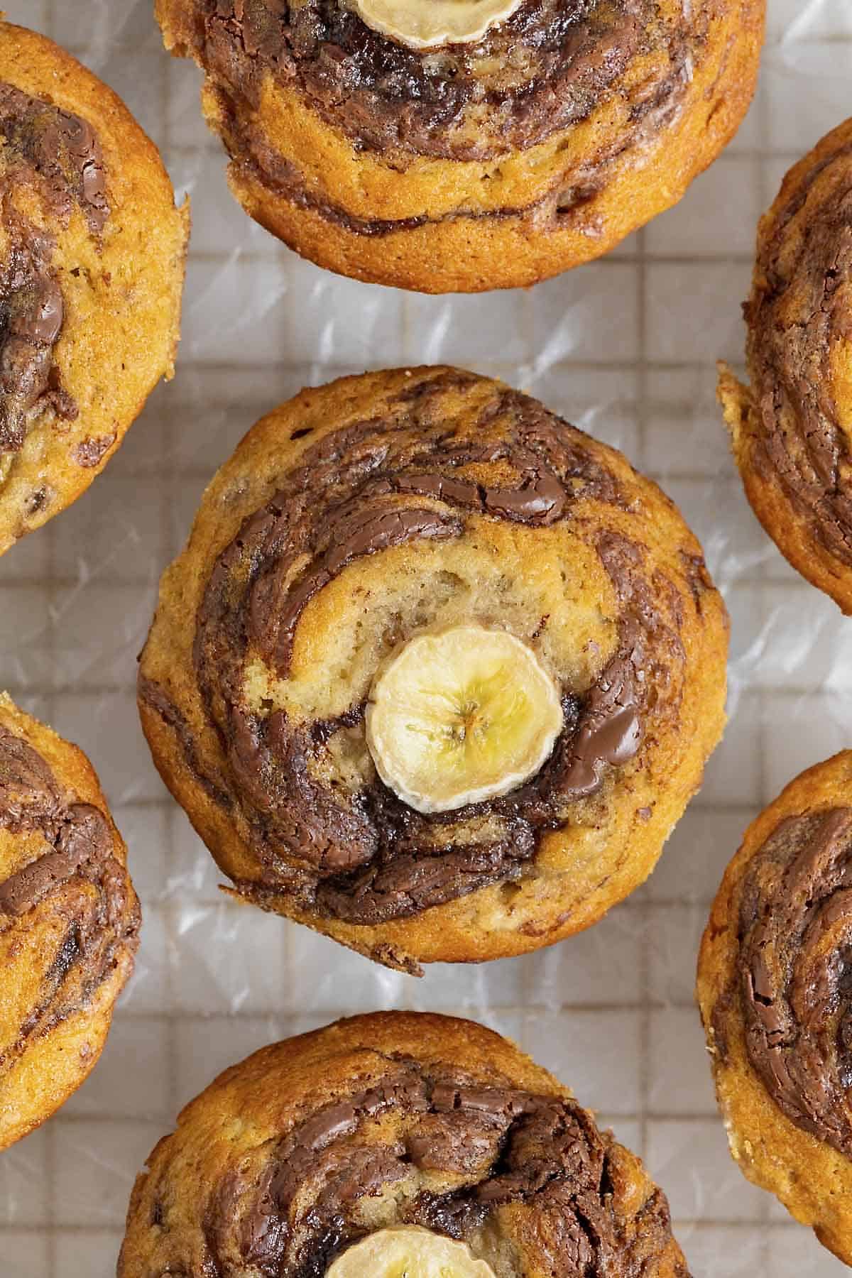 Nutella swirled banana muffins with a slice of banana on top resting on a parchment lined gold tone cooling rack.