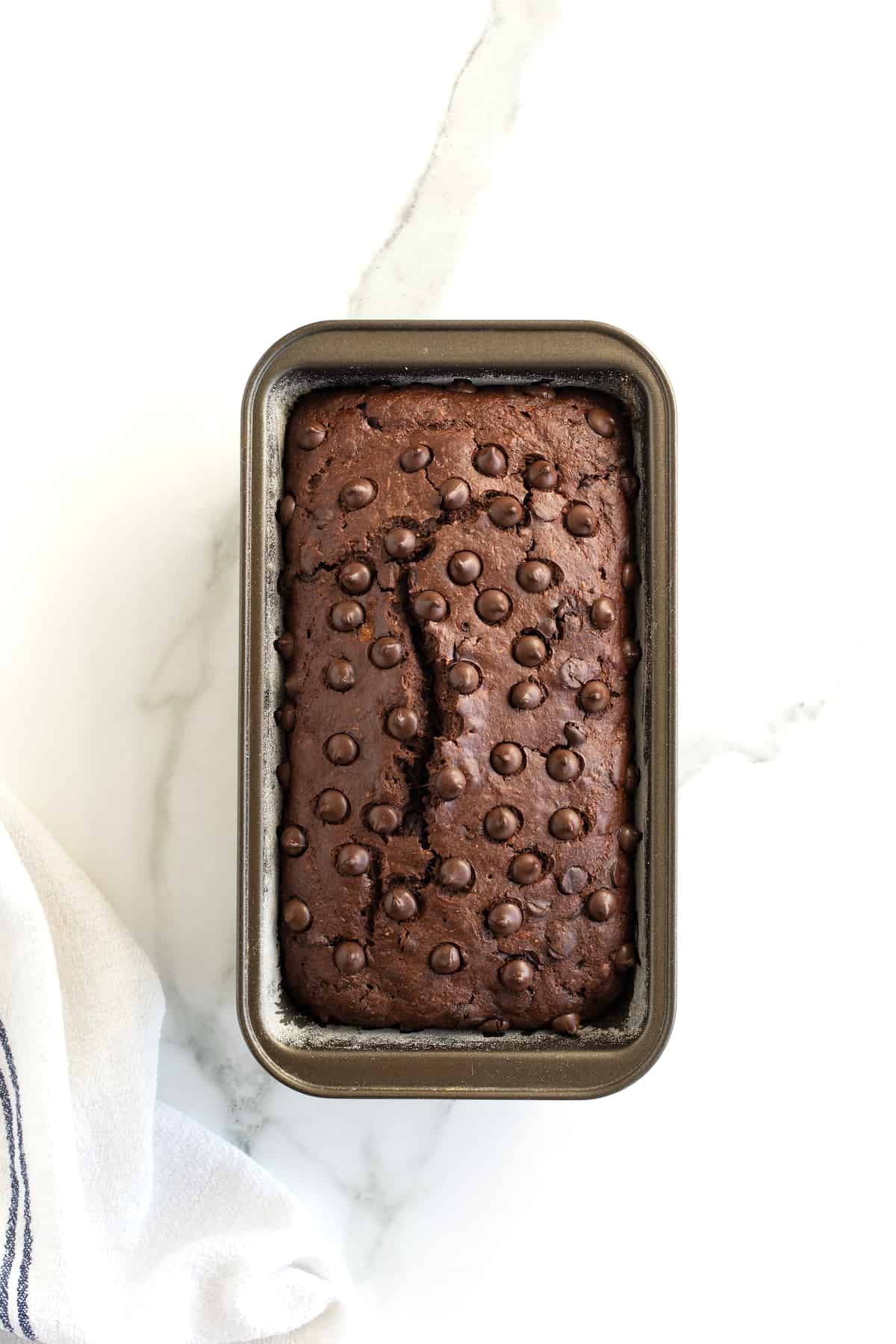 A baked loaf of chocolate banana bread in a metal loaf pan topped with chocolate chips with a crack down the middle.