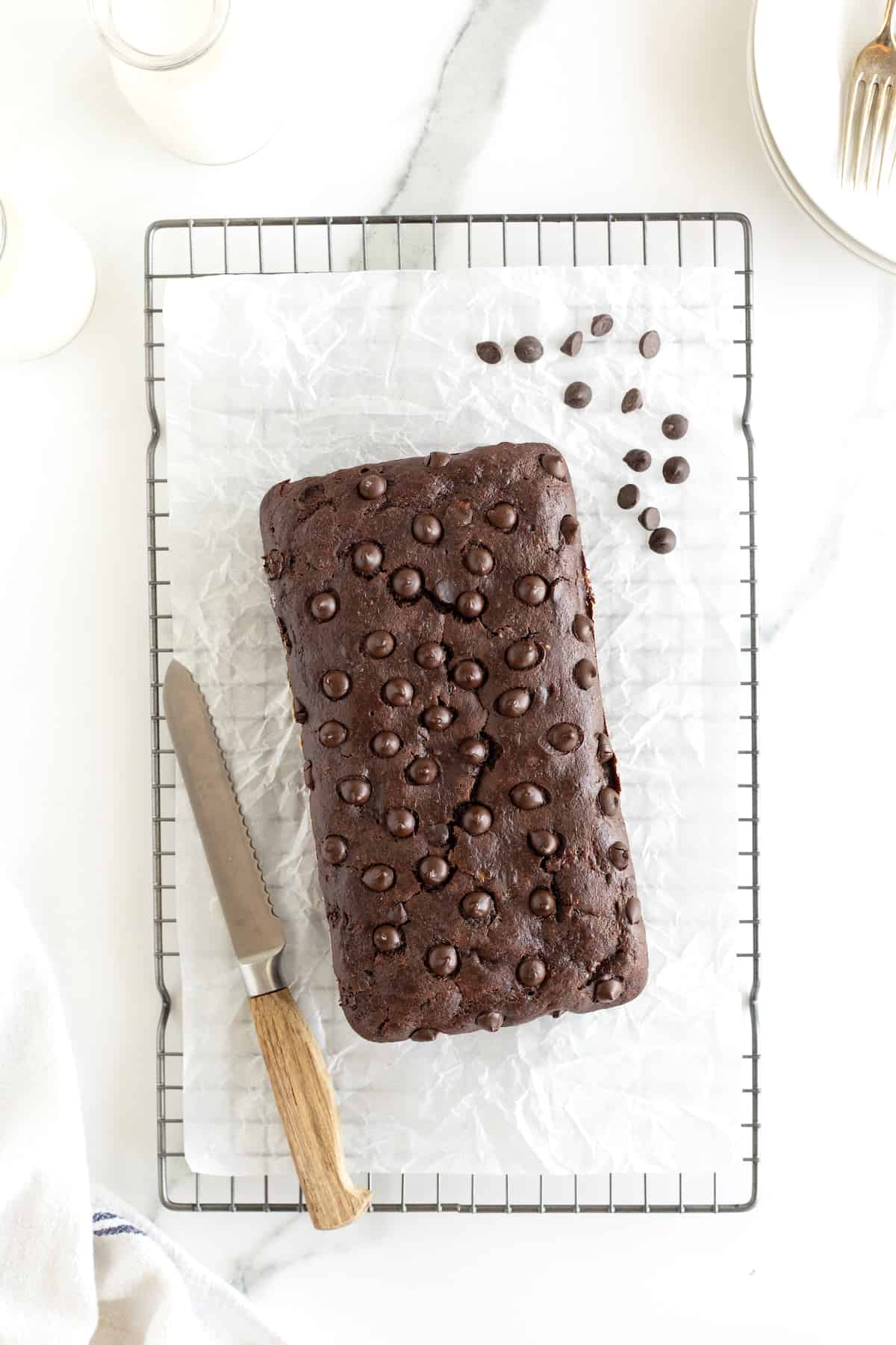 A loaf of chocolate banana bread on a parchment lined cooling rack with scattered chocolate chips and a bread knife.