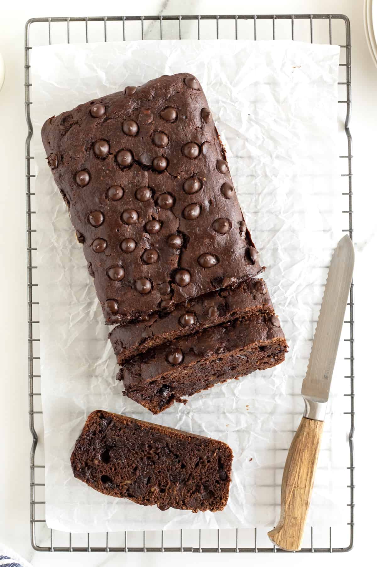 A loaf of chocolate banana bread on a parchment lined cooling rack with a bread knife.