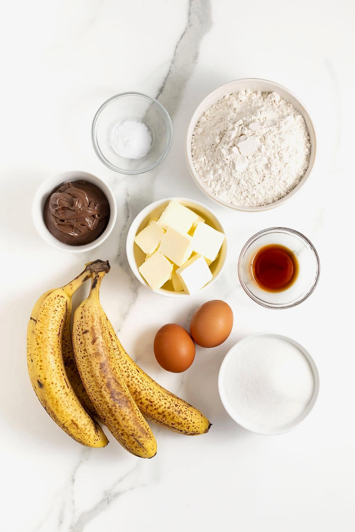 Ingredients to make Nutella swirled banana muffins in small glass dishes on a white marble counter.