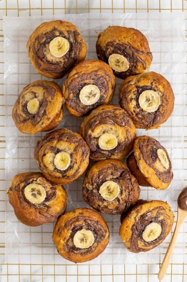 Nutella swirled banana muffins with a slice of banana on top resting on a parchment lined gold tone cooling rack.