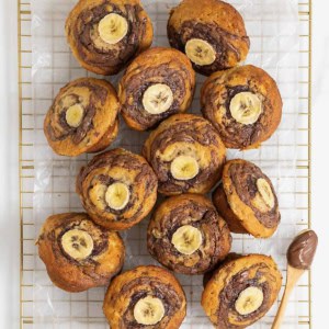 Nutella swirled banana muffins with a slice of banana on top resting on a parchment lined gold tone cooling rack.