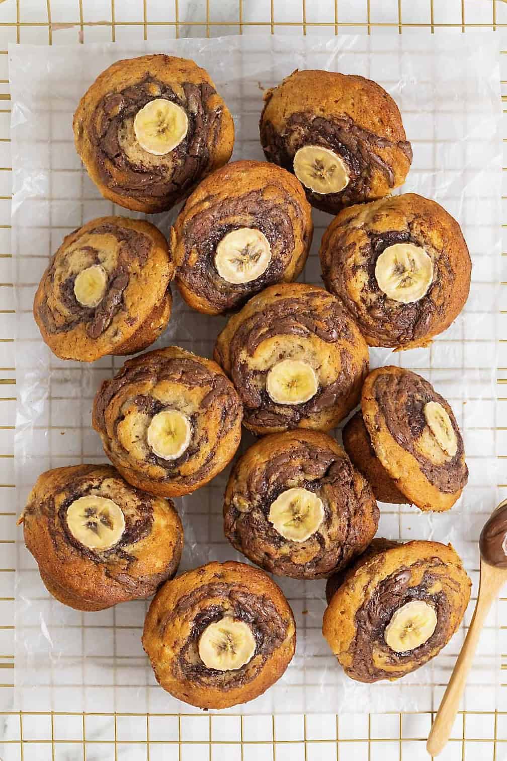 Nutella swirled banana muffins with a slice of banana on top resting on a parchment lined gold tone cooling rack.