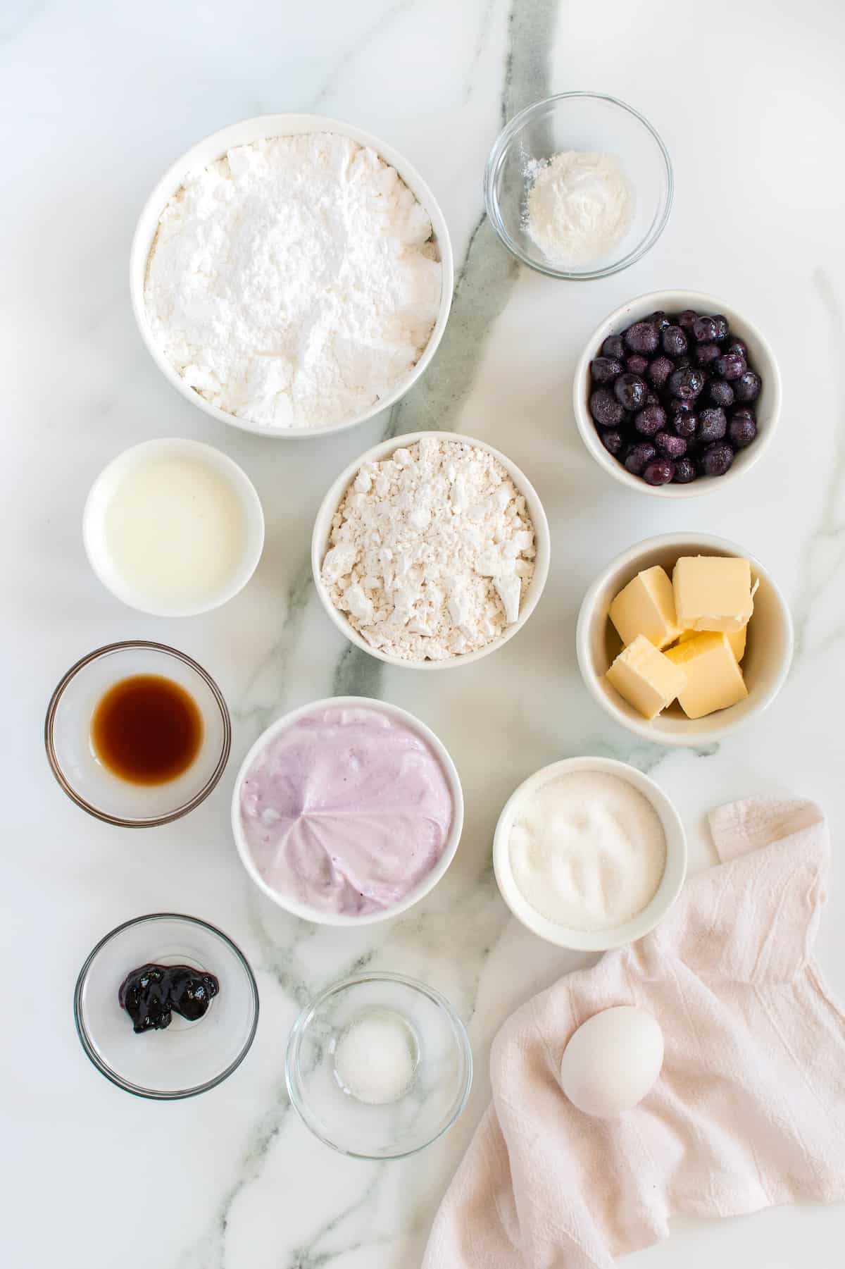 All the ingredients for blueberry fritters in small glass dishes on a white marble counter.