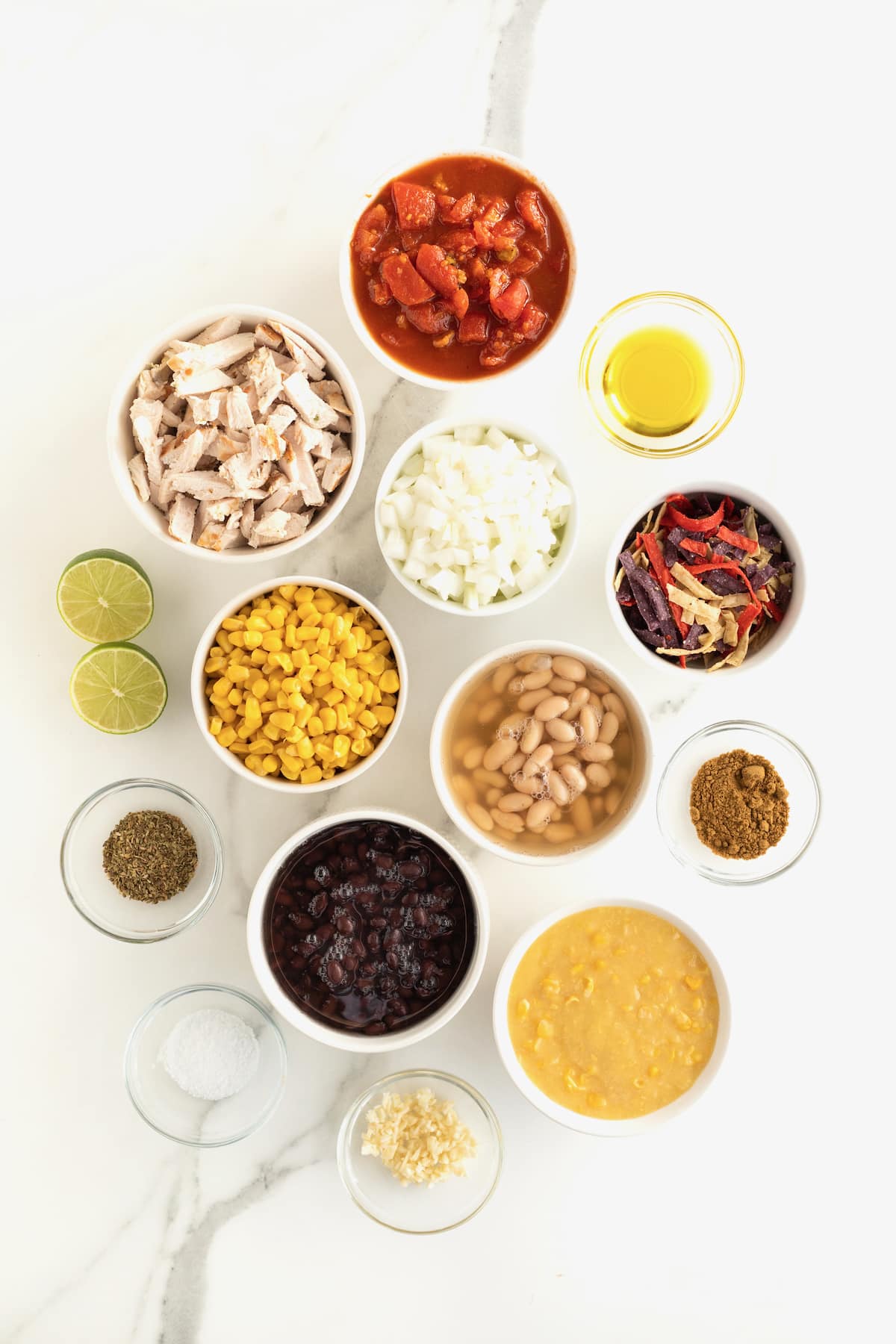 All the ingredients to make chicken tortilla soup in small glass containers on a white counter.