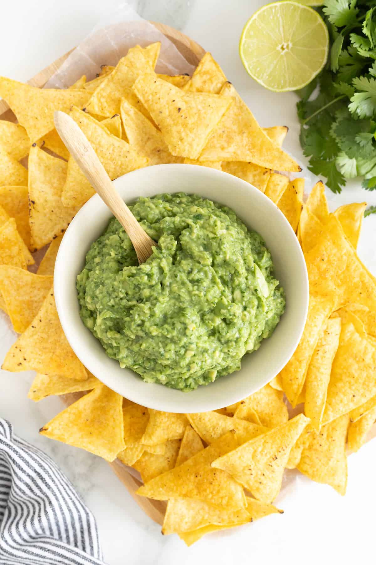 A white bowl of guacamole surrounded by tortilla chips. A small wooden spoon is sticking out of the guacamole.