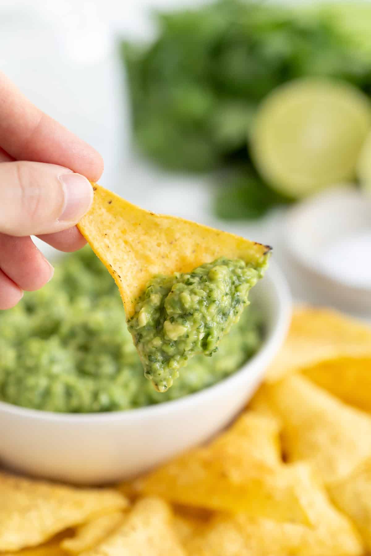 A hand holding a triangular yellow corn tortilla chip with guacamole on it. in the background is a white serving bowl of guacamole.