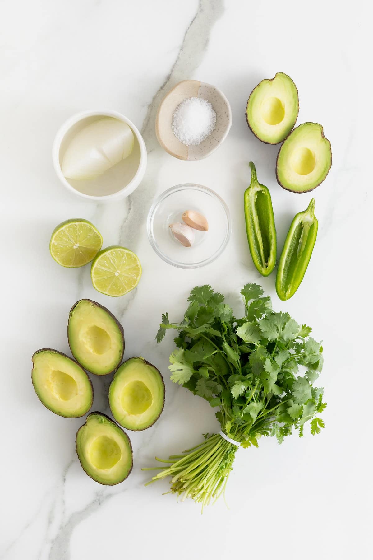 Ingredients to make blender guacamole on a white marble counter.