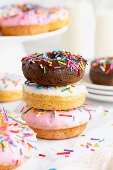 Three donuts in a stack. The top donut is chocolate with chocolate frosting. The middl donut is vanilla with white icing and the bottom donut is vanilla with pink icing. All the donuts have rainbow sprinkles.