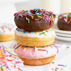 Three donuts in a stack. The top donut is chocolate with chocolate frosting. The middl donut is vanilla with white icing and the bottom donut is vanilla with pink icing. All the donuts have rainbow sprinkles.
