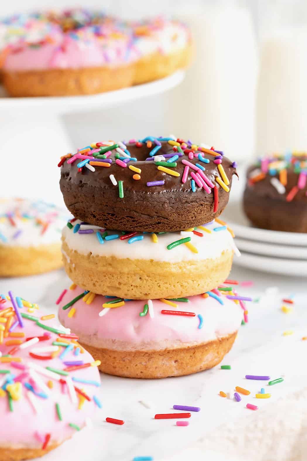 Three donuts in a stack. The top donut is chocolate with chocolate frosting. The middl donut is vanilla with white icing and the bottom donut is vanilla with pink icing. All the donuts have rainbow sprinkles.