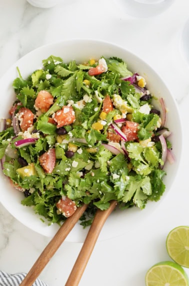 A large white serving bowl of cilantro avocado salad with chopped tomatoes and sliced red onion. There are two wooden salad spoons sticking out of the bowl.