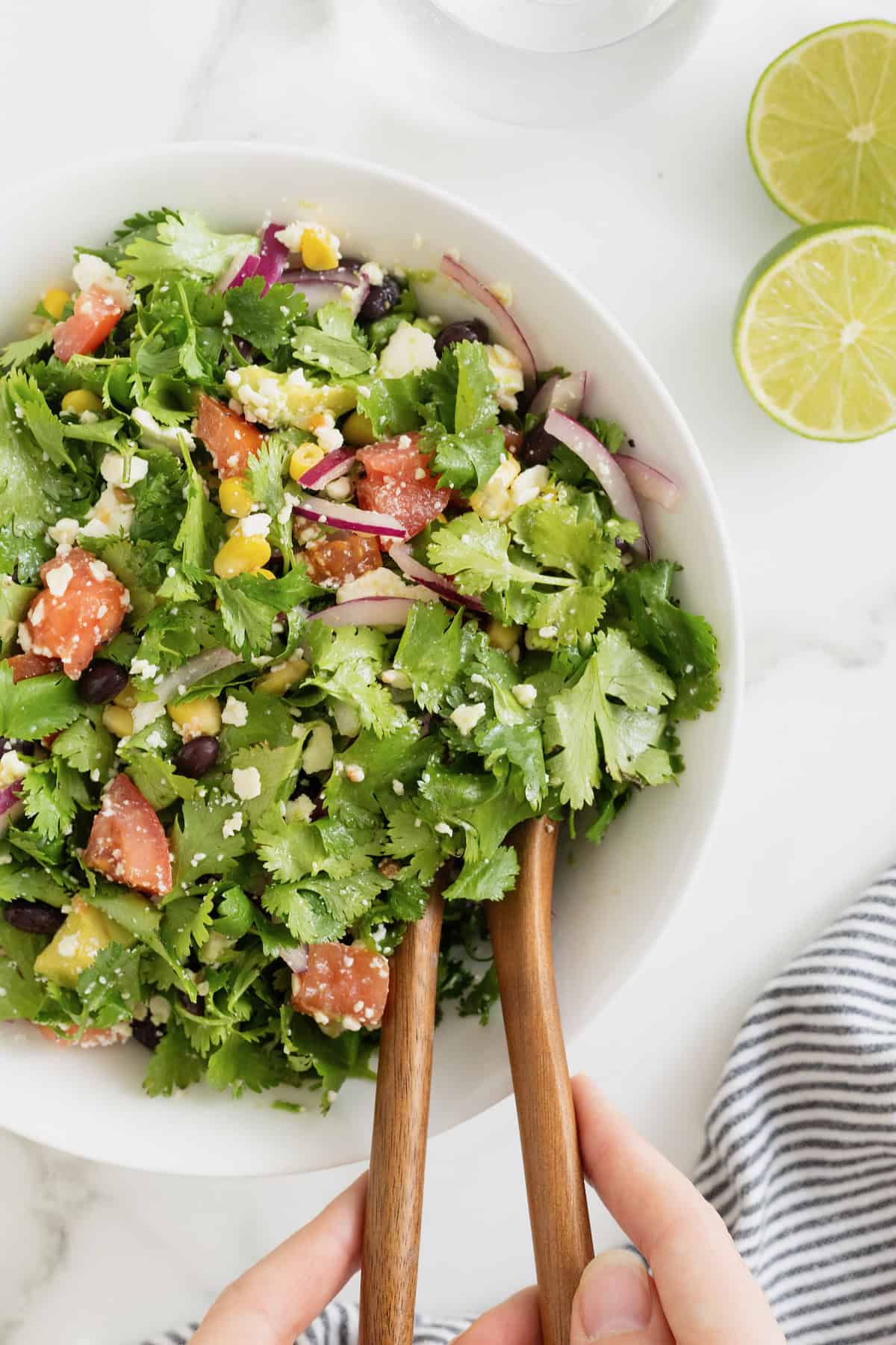A large white serving bowl of cilantro avocado salad with chopped tomatoes and sliced red onion. There are two wooden salad spoons sticking out of the bowl.