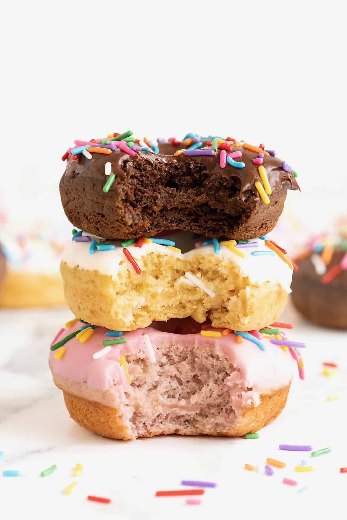Three donuts in a stack. The top donut is chocolate with chocolate frosting. The middl donut is vanilla with white icing and the bottom donut is strawberry with pink icing. All the donuts have rainbow sprinkles and a bite out of them.